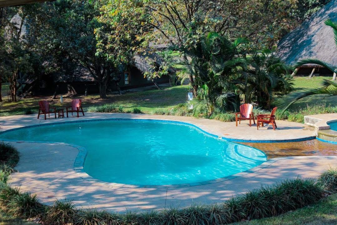 Der geschwungene Außenpool des Njele Country Park bei Ndola, eingebettet in Rasenflächen und alte Bäume, dahinter strohgedeckte Chalet-Dächer